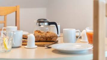 A breakfast table with a coffee pot, a loaf of bread, a glass of orange juice, and white cups.