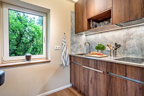A modern kitchen with wooden cabinets, a marble countertop, a window with a view of greenery, and a sink with utensils.