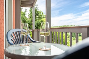 A glass table with two wine glasses on coasters, a blue chair, and a view of greenery through a balcony railing.
