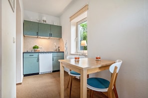 A compact kitchen with a dining area, featuring a wooden table and chairs, a small refrigerator, and a window with a view of greenery. - Studio Apartment 'Mohnrot - Fulgenhaus' with Balcony (Wittenbeck-Klein Bollhagen)