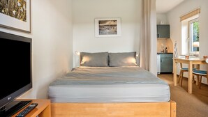 Un dormitorio con cama, televisor, control remoto, un cuadro en la pared y una pequeña cocina.