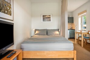 A bedroom with a bed, a TV, a remote, a framed picture on the wall, and a kitchenette area.