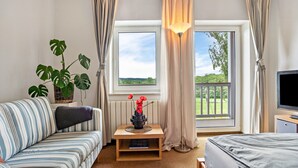 Una habitación con sofá, mesita de noche con lámpara, televisor y ventana con cortinas.
