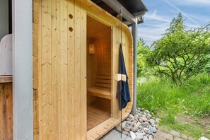 A wooden sauna with a bench inside, a towel hanging on the door, and a stone pathway outside.