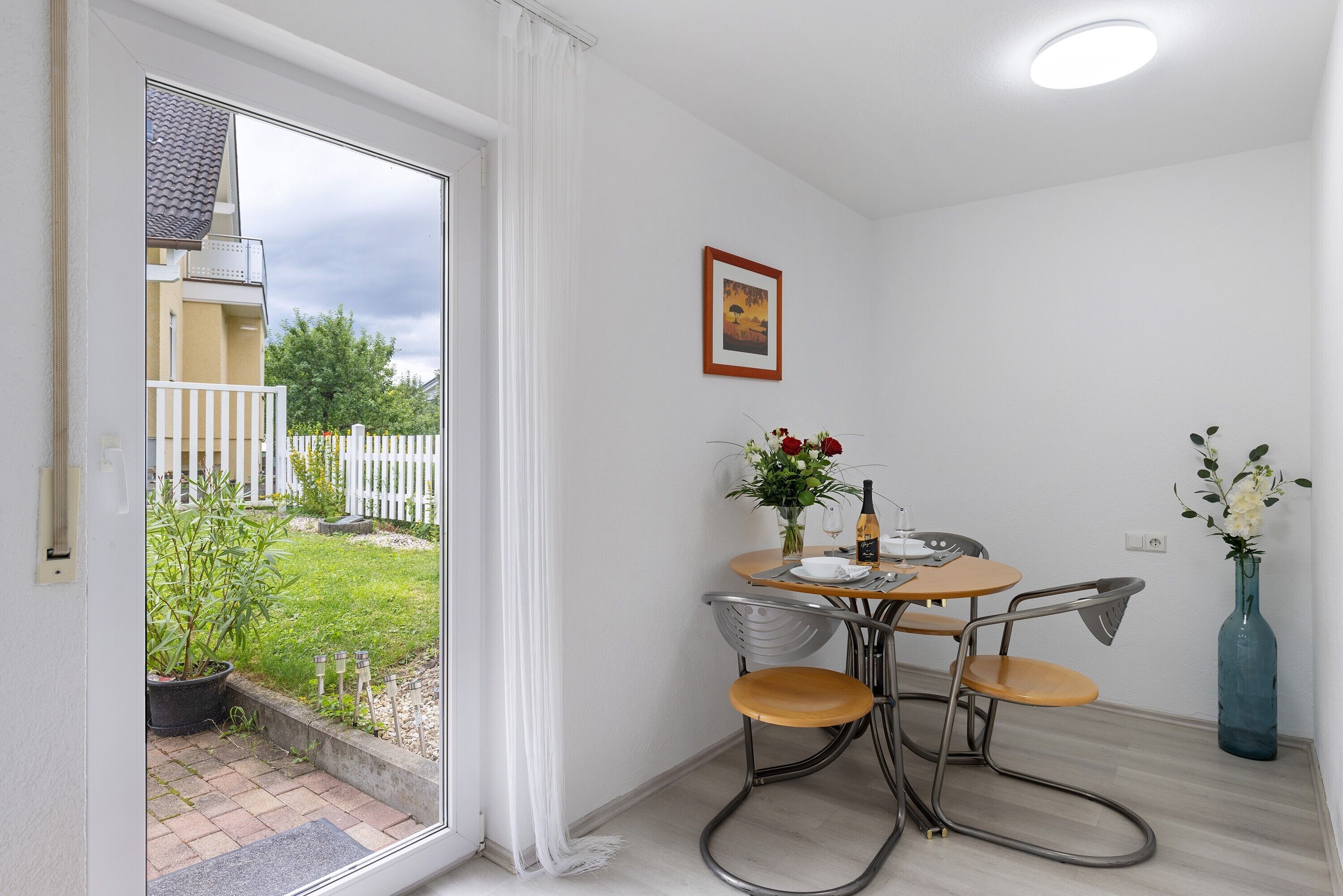 A room with a sliding glass door leading to a balcony, a small round table with chairs, a vase with flowers, and a framed picture on the wall.