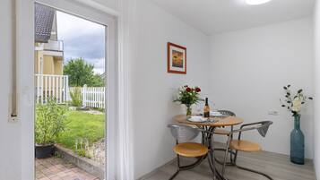 A room with a sliding glass door leading to a balcony, a small round table with chairs, a vase with flowers, and a framed picture on the wall.