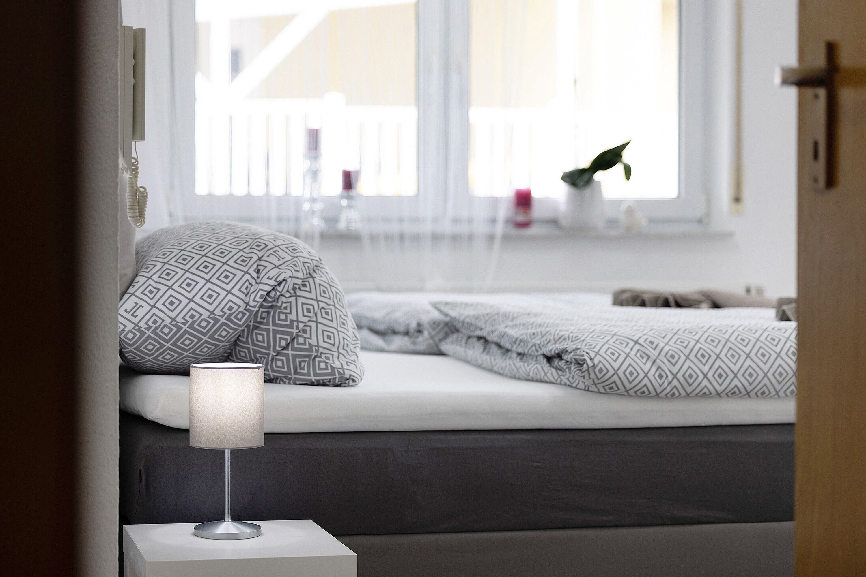 Et soveværelse med en seng, mønstrede puder, en sengetavlelampe og et vindue med udsigt til en balkon.