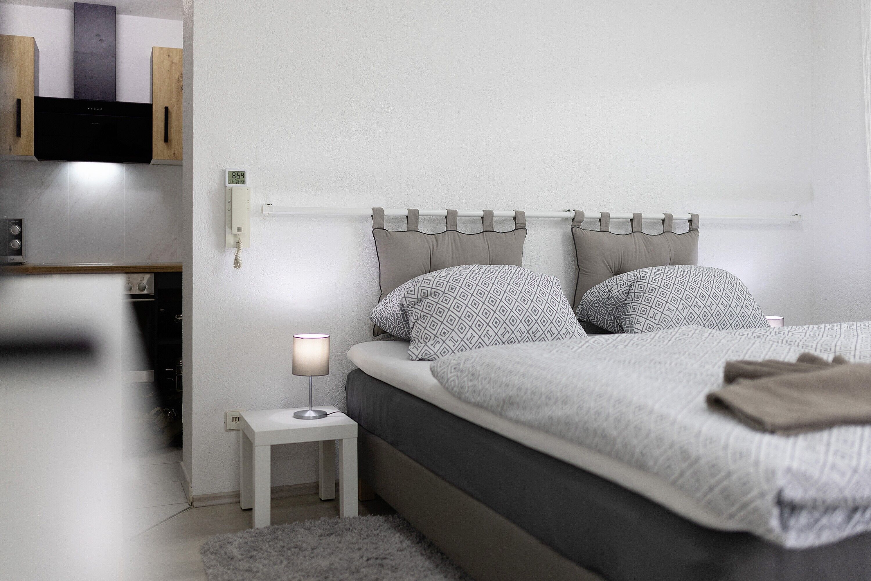 Et soveværelse med seng, sengebord, lampe og et vægmonteret termostat.