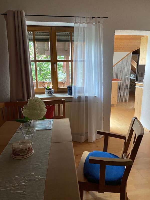A dining area with a table, chairs, a vase with flowers, a window with curtains, and a view of an outdoor deck.