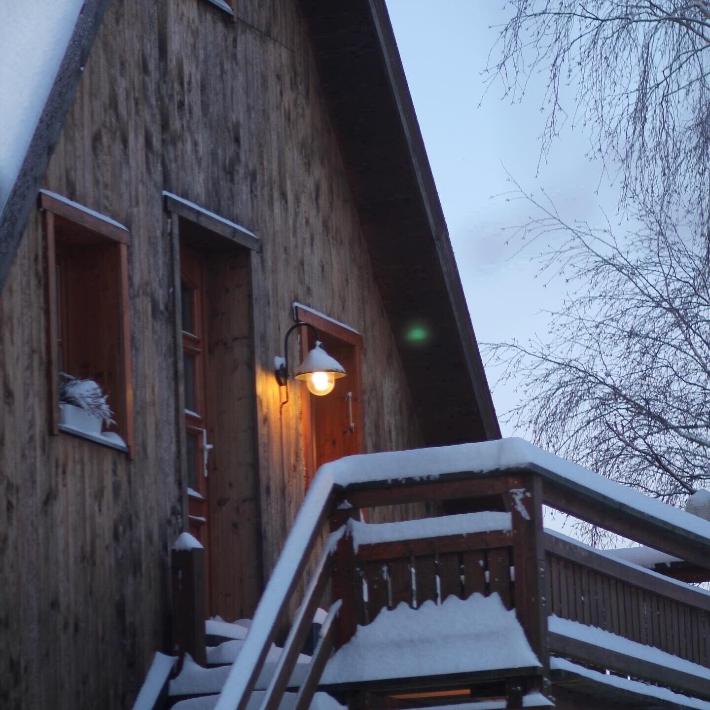 A wooden building with a staircase leading to a balcony, snow on the roof, and a lit exterior light.