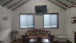 A living room with a high ceiling, a sofa, a coffee table, a television, and two windows with blinds. - Windy Ridge Cabin
Babb/St.Marys (Babb)