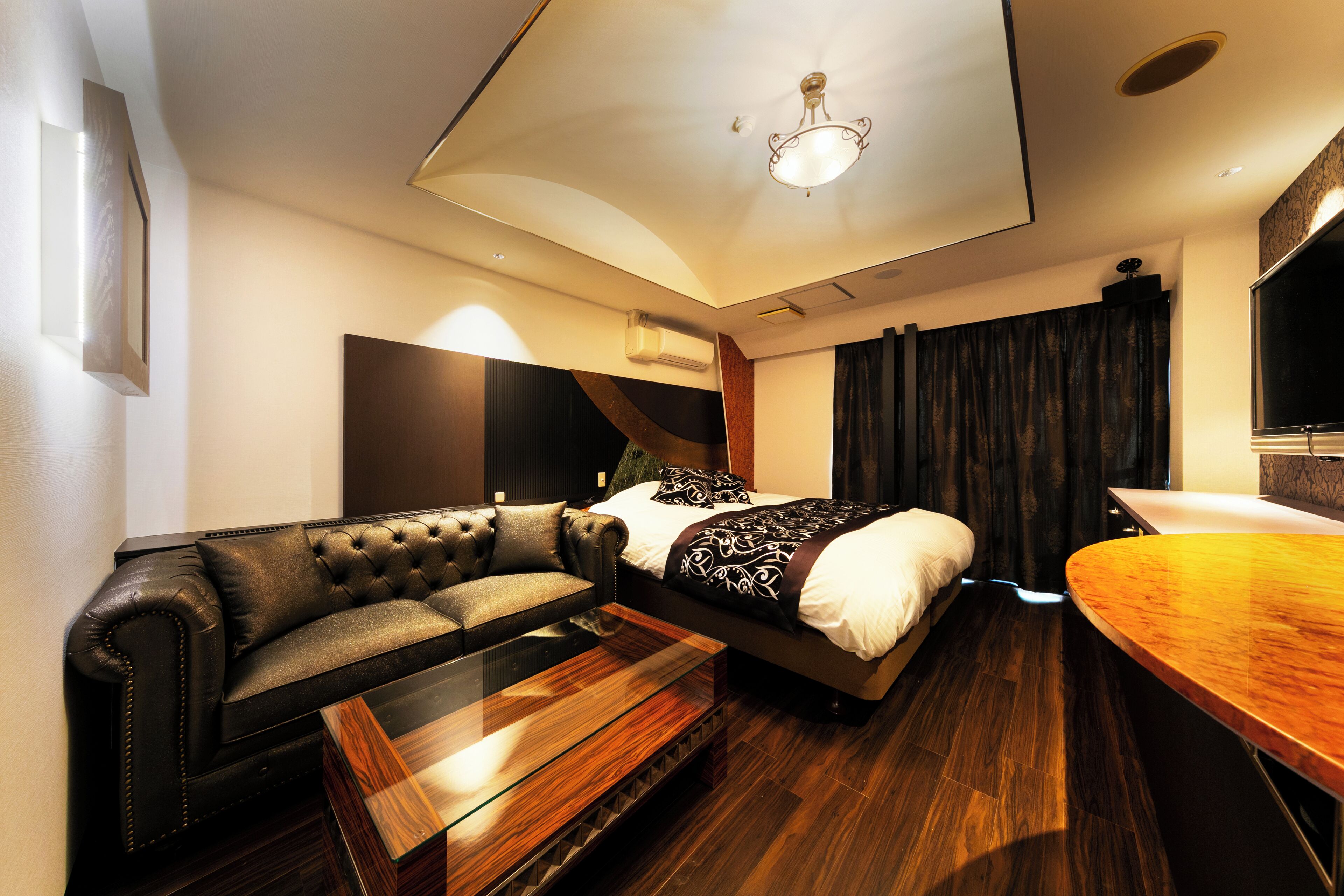 A modern hotel room with a bed, a leather sofa, a wooden coffee table, and a curved wooden desk.