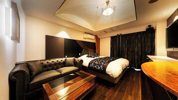 A modern hotel room with a bed, a leather sofa, a wooden coffee table, and a curved wooden desk.