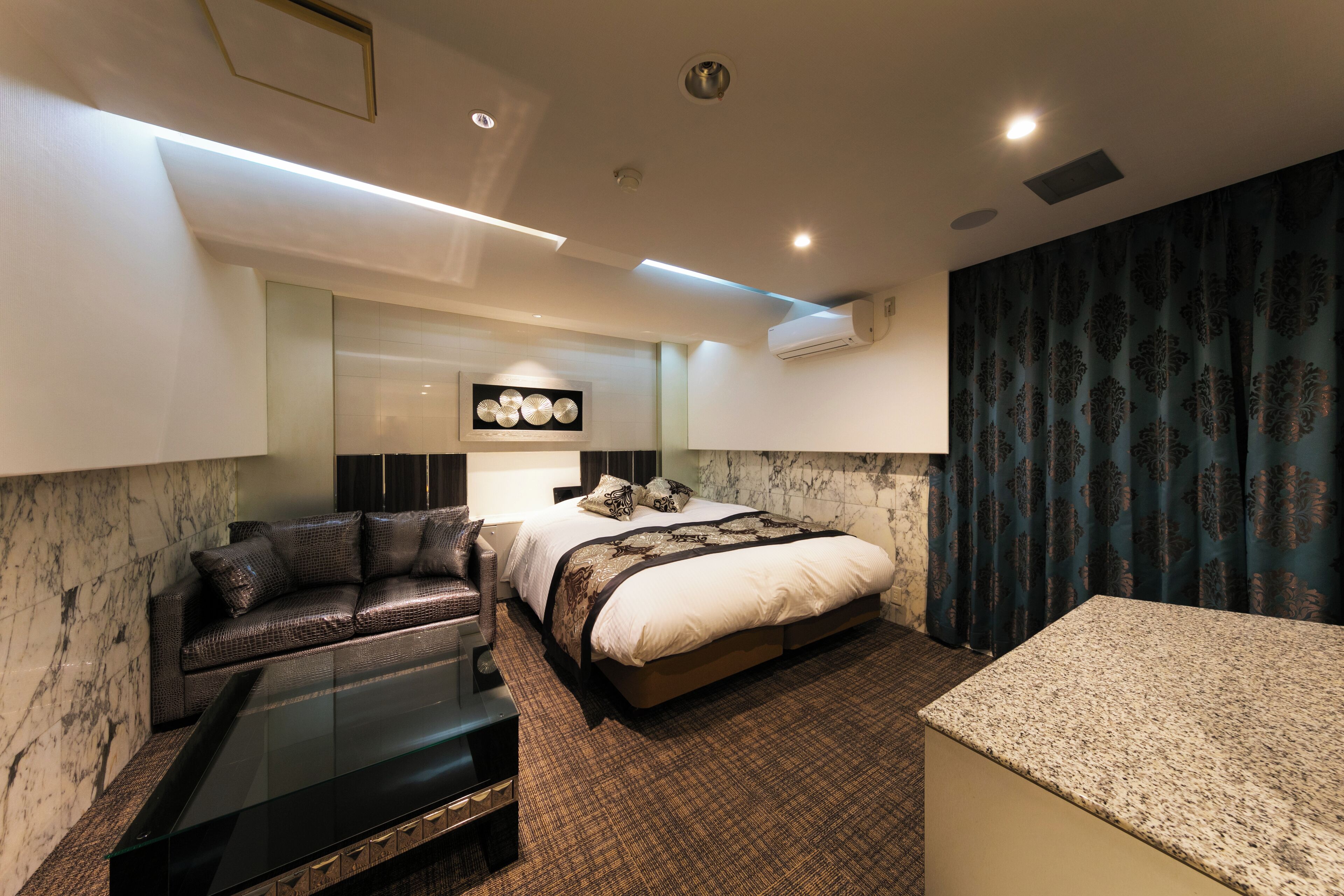 A modern hotel room with a bed, a sofa, a glass coffee table, and a marble accent wall.