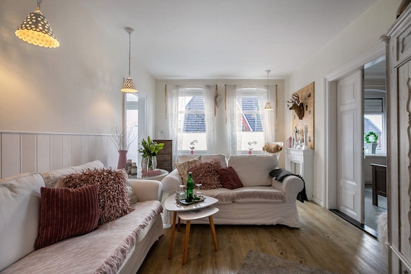 A cozy living room with two sofas, a coffee table, and a dining table. There are windows with curtains, a wall-mounted deer head decoration, and a door leading to another room.