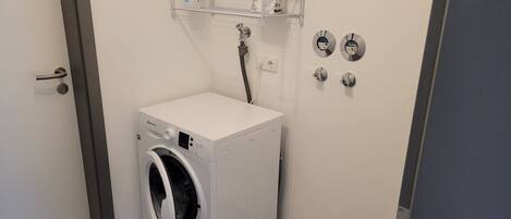 A compact laundry room with a front-loading washing machine, a shelf with toiletries, and a wall-mounted hand dryer.