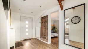 Un pasillo con piso de madera, paredes blancas, una puerta de madera, un espejo con un reloj y una cama al fondo.