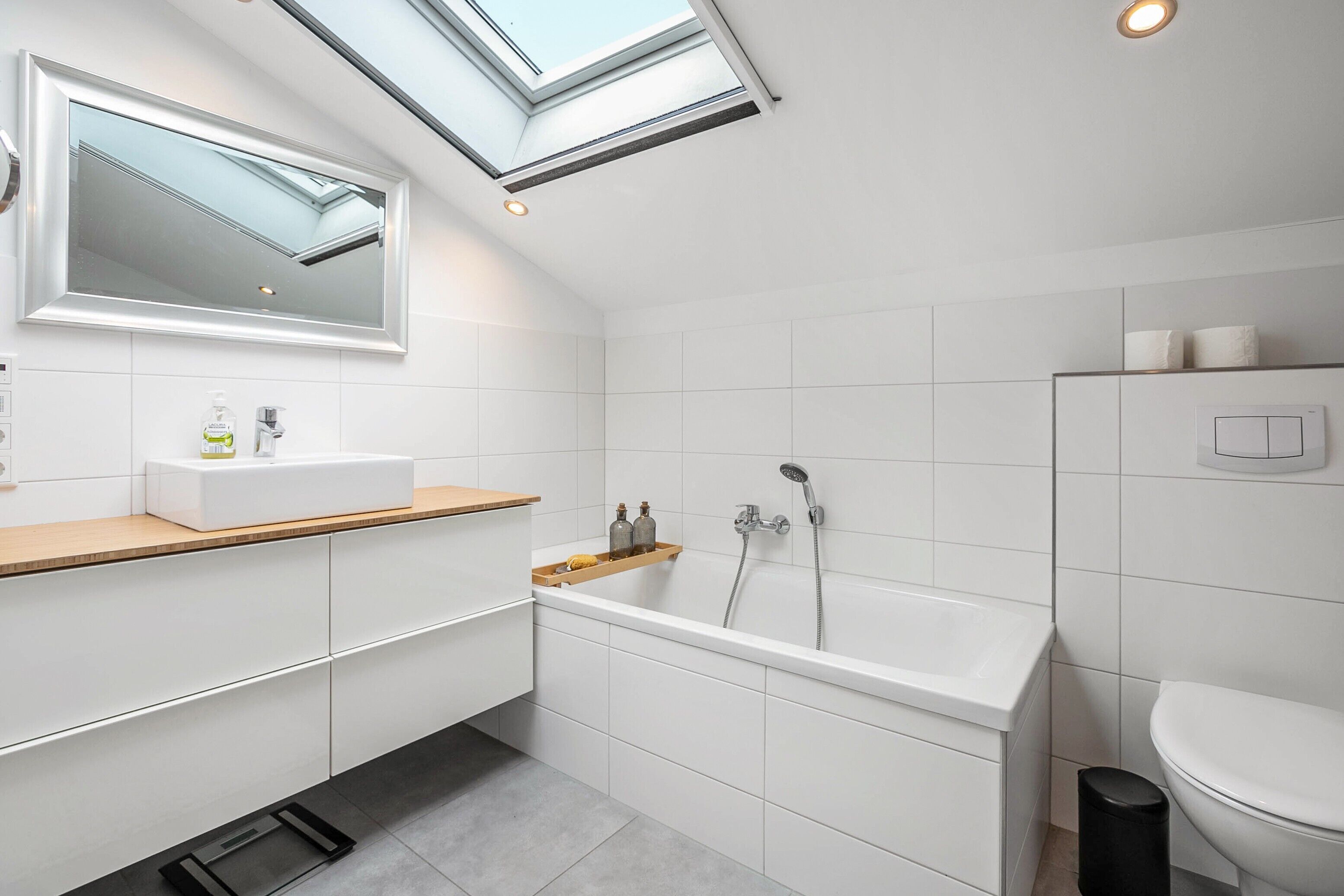 Une salle de bain moderne dotée d’un puits de lumière, d’une baignoire, de toilettes et d’un lavabo avec un plan de travail en bois.