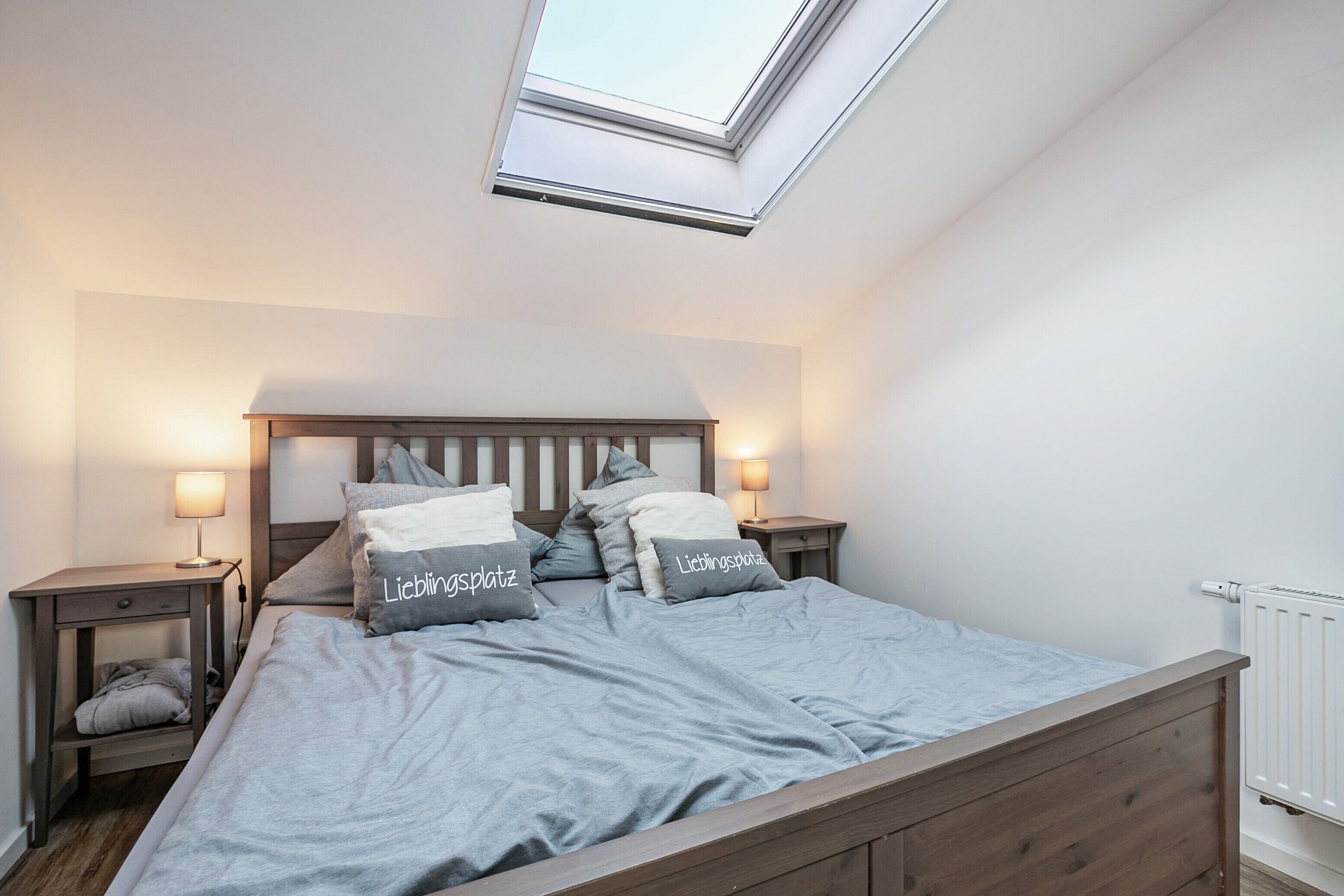 Ein Schlafzimmer mit Oberlicht, einem Bett mit grauer Bettwäsche, zwei Nachttischen mit Lampen und einer Heizung.
