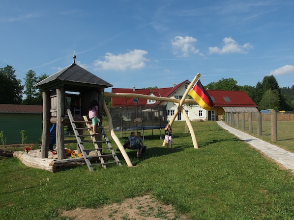 Une aire de jeux avec une structure en bois, un trampoline et un mât sur lequel flotte un drapeau.
