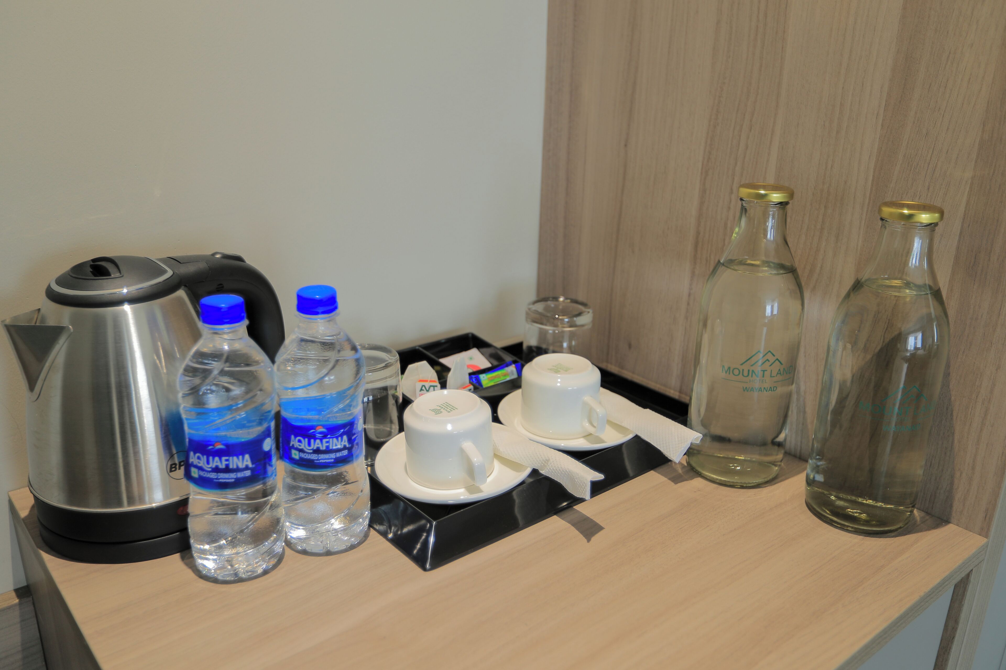Un plateau de petit-déjeuner dans une chambre d’hôtel, avec une bouilloire, deux bouteilles d’eau, deux tasses blanches, une serviette en papier et deux bouteilles en verre vides.