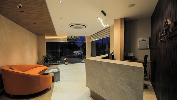 A modern hotel reception area with a curved orange seating area, a marble reception desk, and a view of the cityscape through large windows. - Mount Land (Sulthan Bathery)