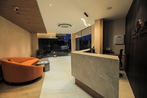 Eine moderne Hotelrezeption mit einer gebogenen orangefarbenen Sitzgruppe, einem Marmor-Rezeptionsschalter und Blick auf die Stadt durch große Fenster.
