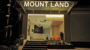 The exterior of a hotel named "Mount Land" with a modern design, featuring a reception desk, a staircase leading to the entrance, and potted plants around the entrance area. - Mount Land (Sulthan Bathery)