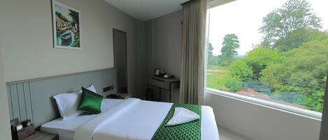 Habitación de hotel con cama, mesita de noche, escritorio, un cuadro y ventana con vistas a zonas verdes.