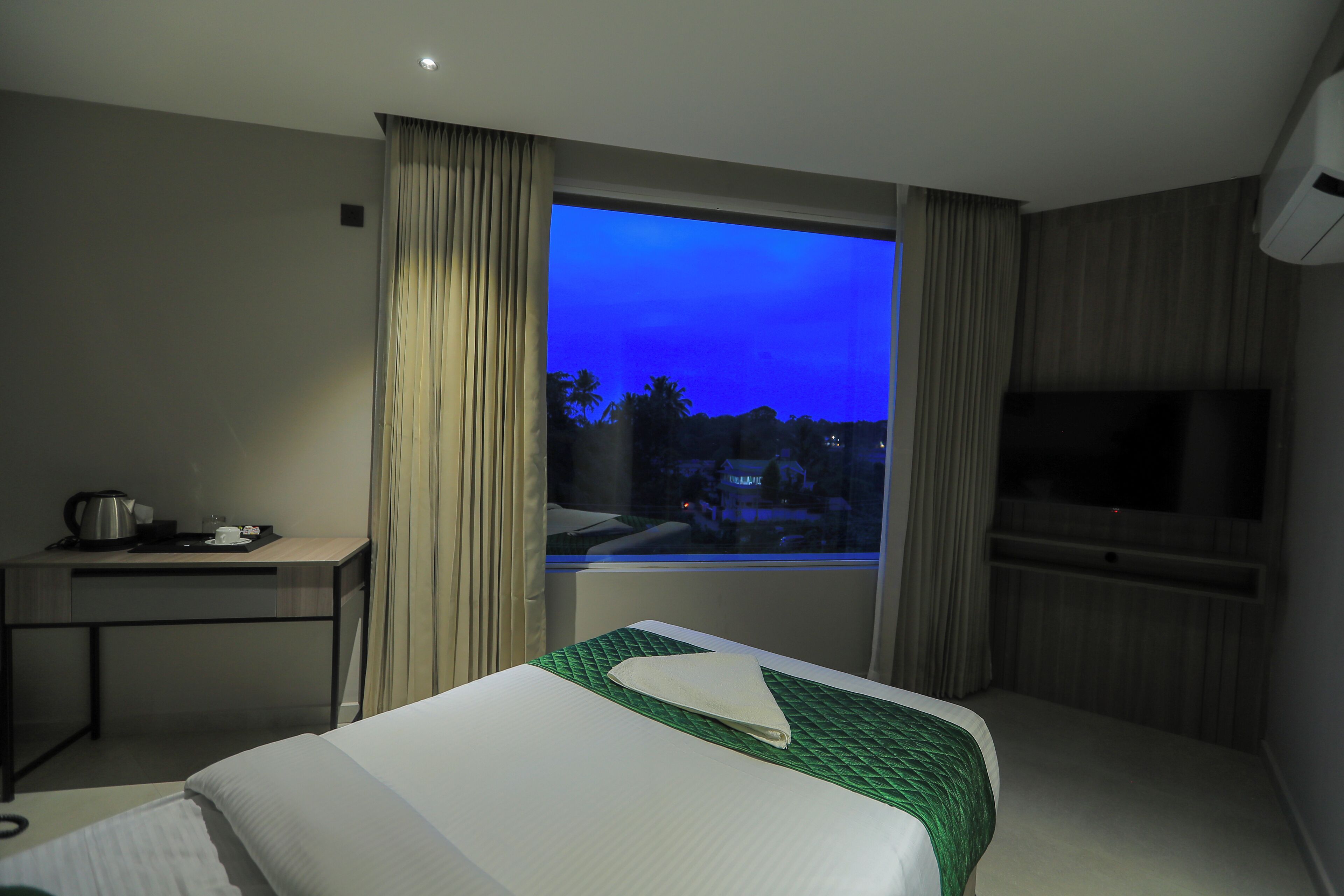 Habitación de hotel con cama, escritorio con hervidor, televisor y ventana con cortinas.