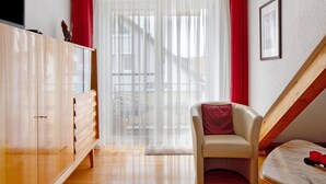 A bedroom with a wooden wardrobe, a chair, a bed, a window with sheer curtains, and a staircase.