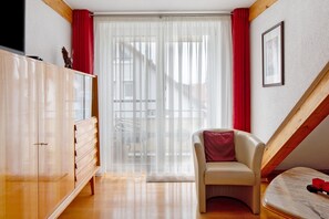 A bedroom with a wooden wardrobe, a chair, a bed, a window with sheer curtains, and a staircase.