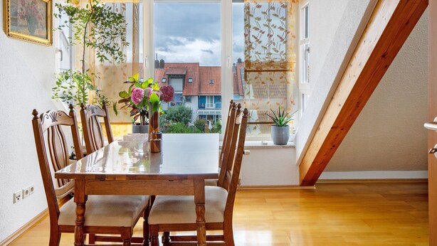 A dining area with a wooden table and chairs, a vase with flowers, and a view of buildings through a window.