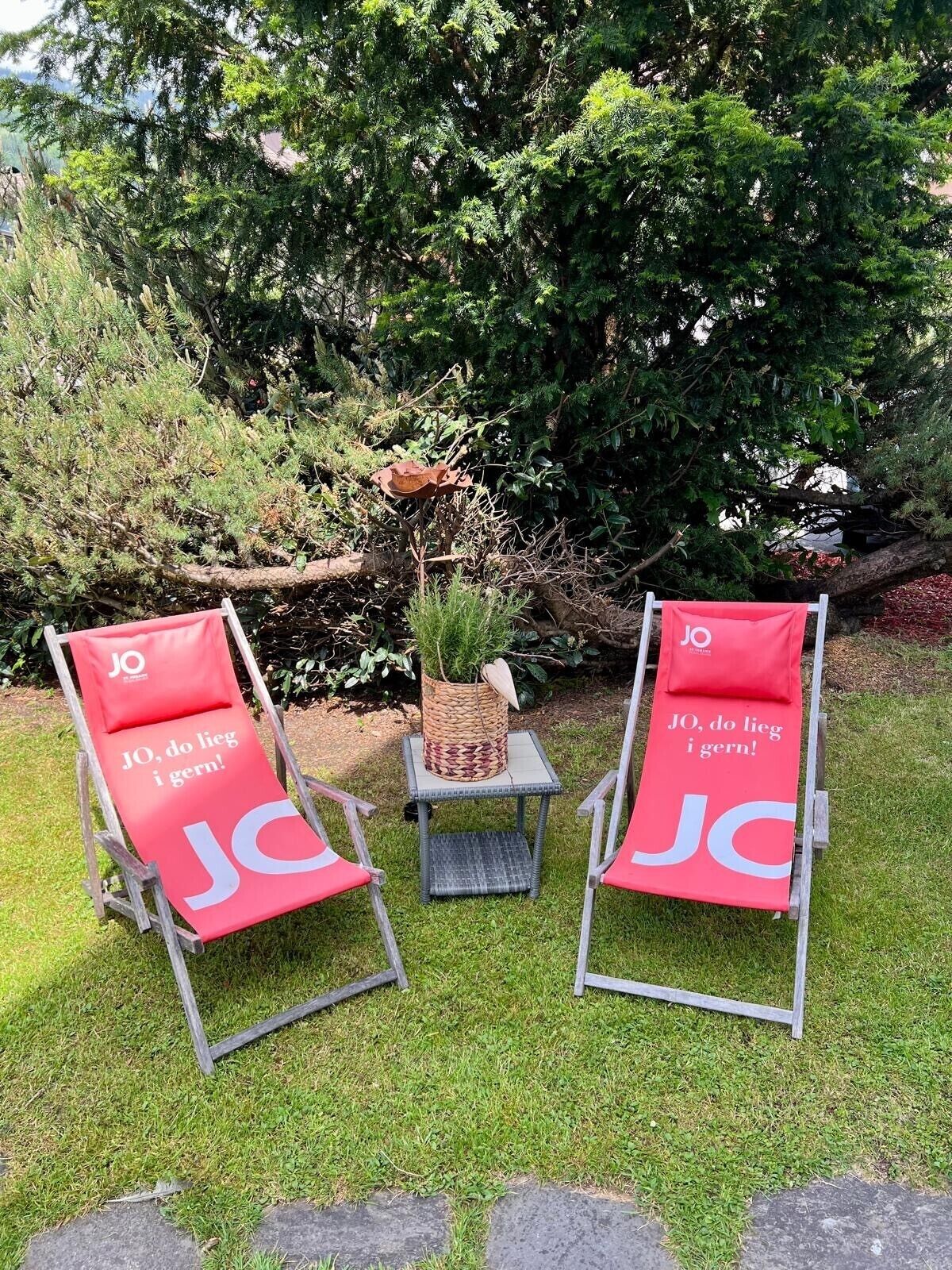 Two red lounge chairs with 'JO' branding, a woven basket, and a small table in a grassy garden with dense foliage in the background.