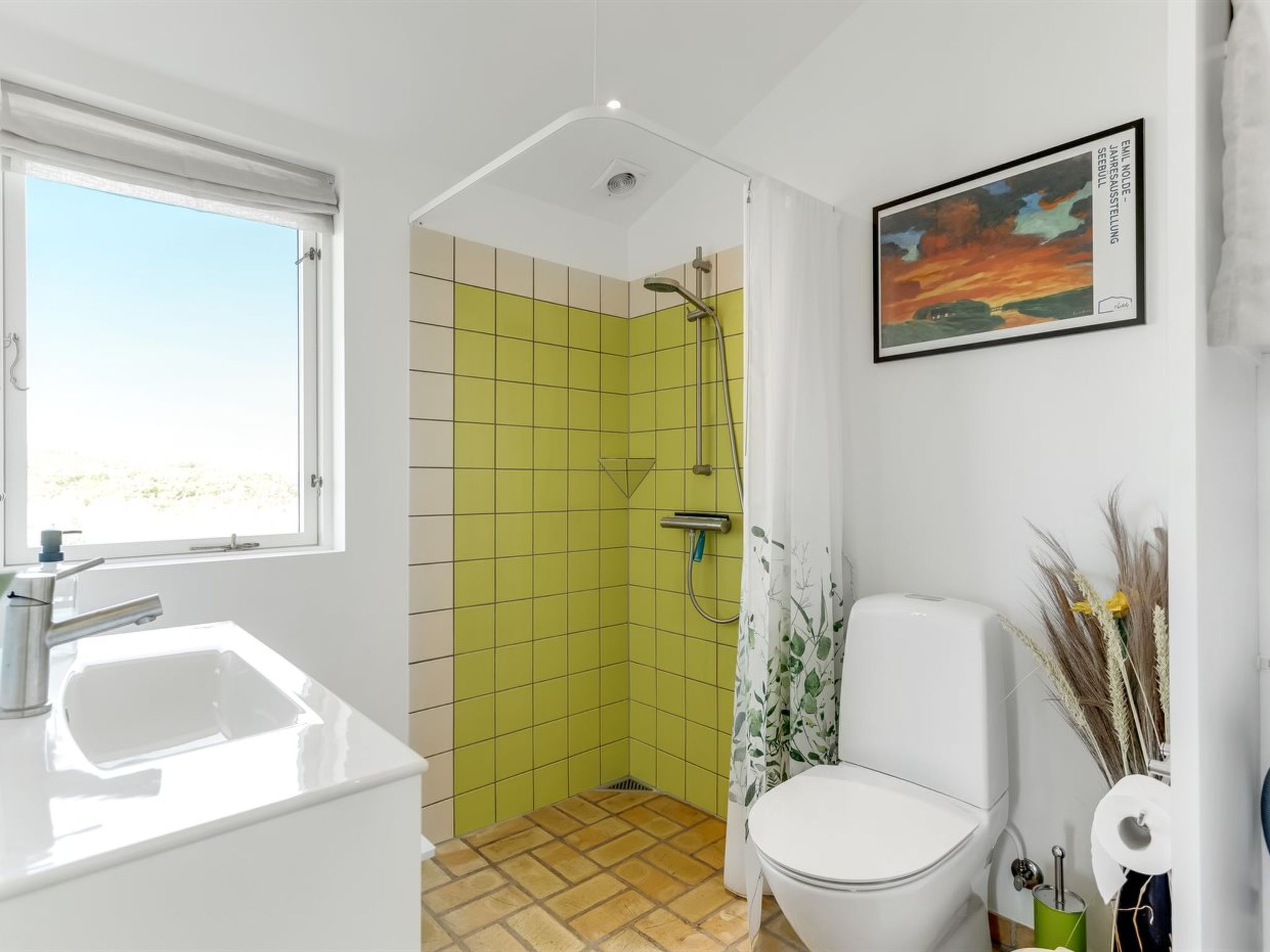 Une salle de bain avec une fenêtre, une douche aux carreaux verts, des toilettes et un tableau encadré au mur.