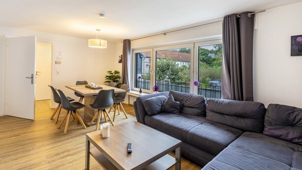 A modern living room with a sofa, dining table, chairs, and a large window with curtains. - 'Apartment by the Park' with Mountain View and Balcony (Willingen (Upland))