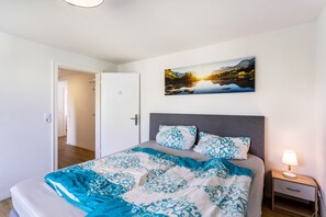 A bedroom with a bed, a nightstand with a lamp, a door, and a wall-mounted picture of a landscape.