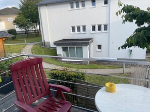 A balcony with a red chair and a small table, overlooking a white building with multiple windows and a pathway leading to it.