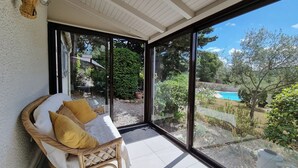 A wicker sofa with cushions, a glass door leading to a garden with a pool, and a ceiling with wooden beams.