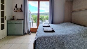A bedroom with a bed, a small cabinet, a window with curtains, and a view of greenery outside.