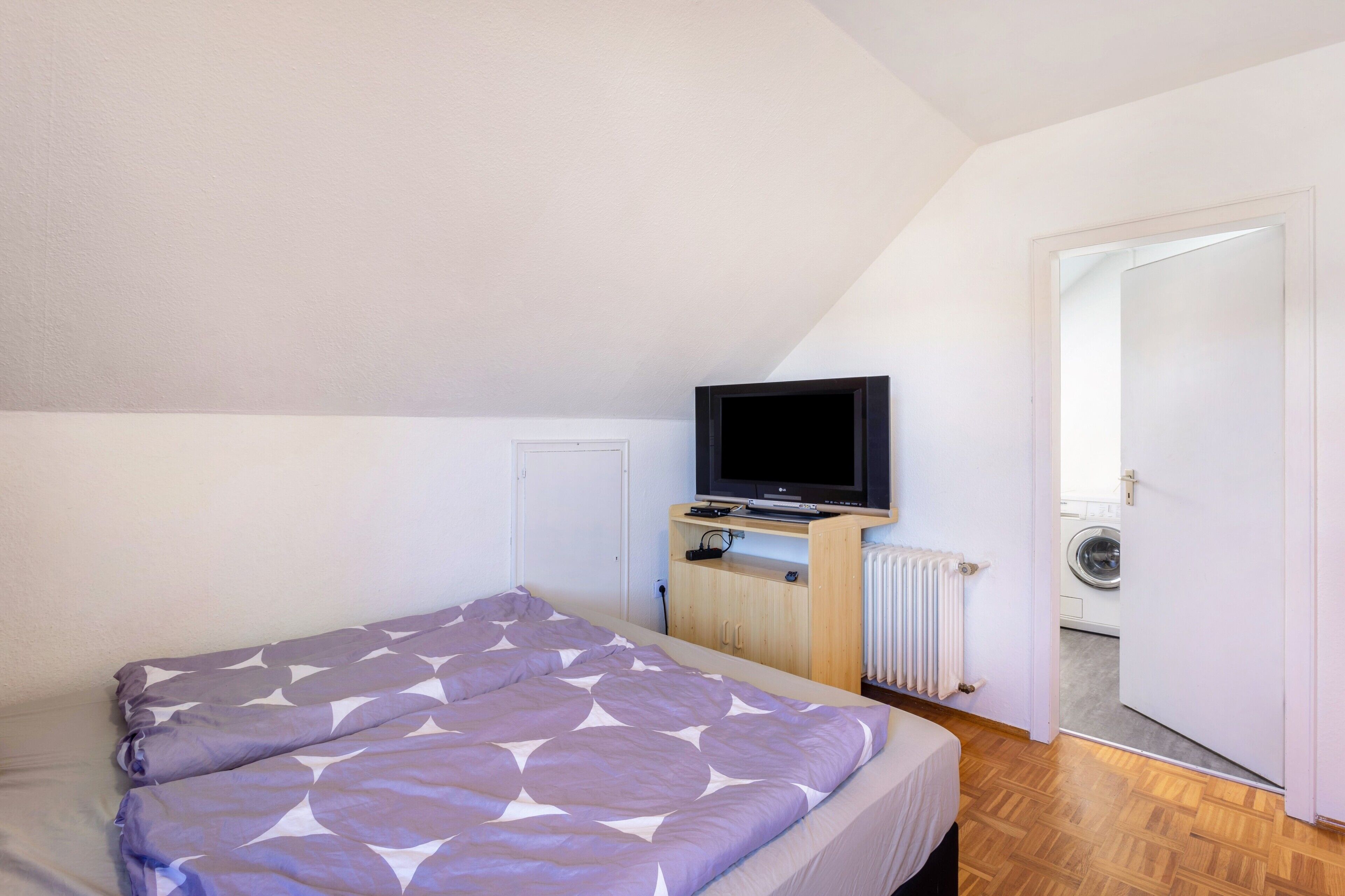 A bedroom with a bed, a television on a stand, a radiator, and a door leading to a laundry area.