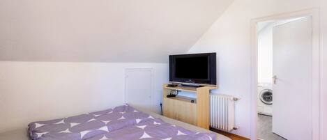 A bedroom with a bed, a television on a stand, a radiator, and a door leading to a laundry area.
