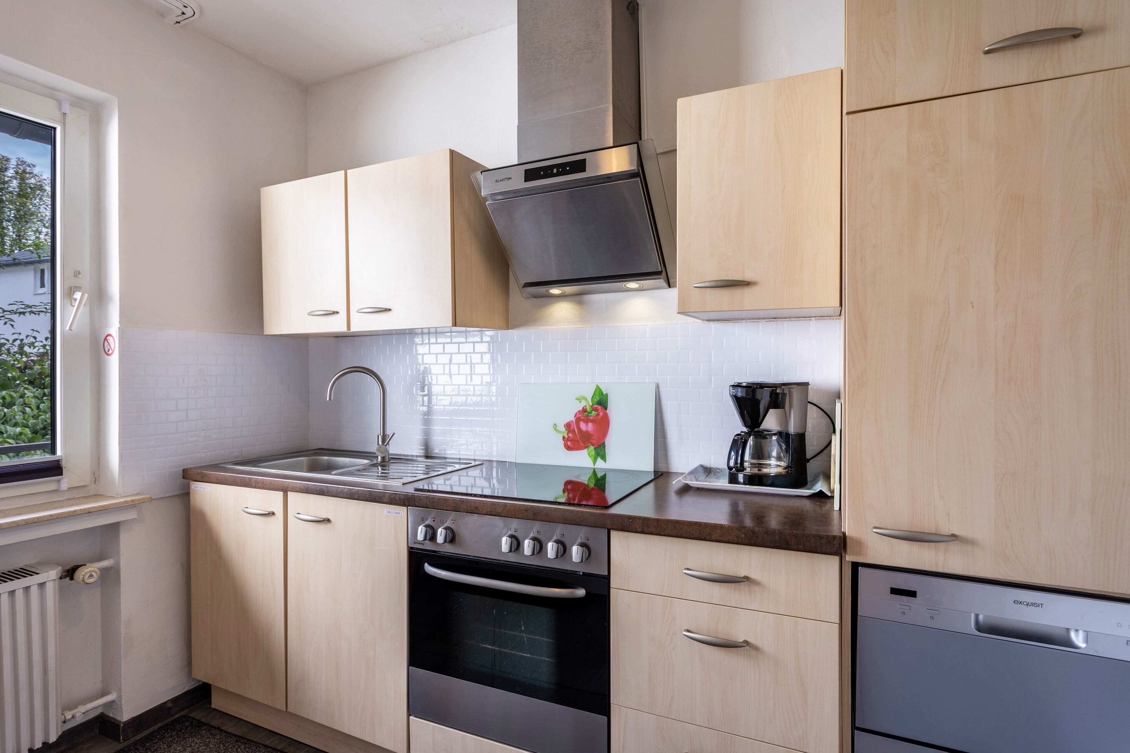 A modern kitchen with light wood cabinets, stainless steel appliances, and a window providing natural light.