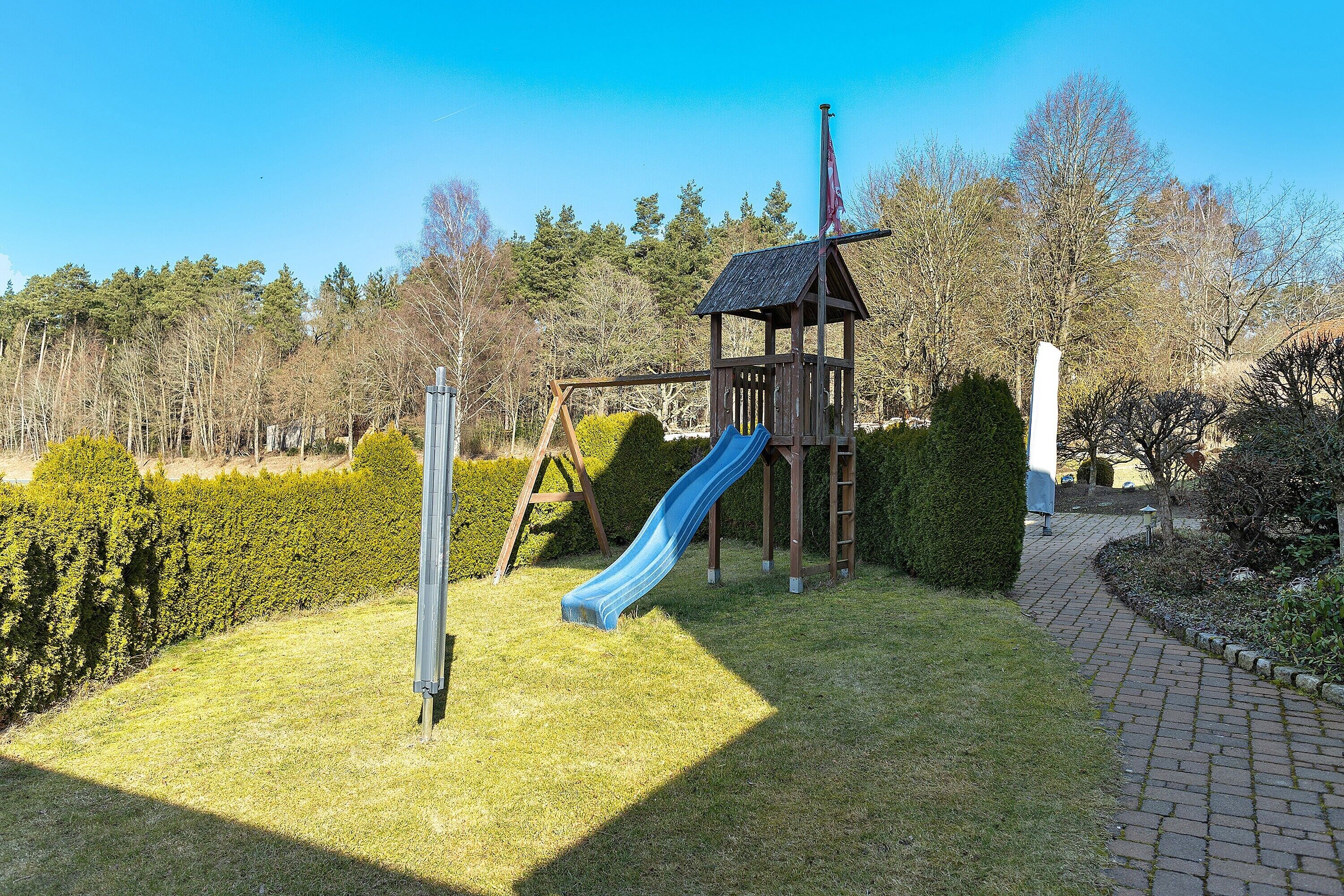 Ein Spielplatz mit Rutsche und Schaukeln, umgeben von Hecken und Bäumen.