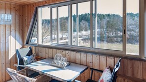 Ein Zimmer mit Holzwänden, einem großen Fenster, einem Tisch mit dekorativen Objekten und einem Korbstuhl.