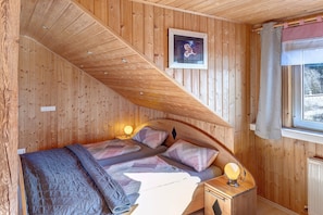 Ein Zimmer mit Holzwänden, einem Bett, einem Nachttischchen mit Lampe, einem gerahmten Bild an der Wand und einem Fenster mit Vorhang.