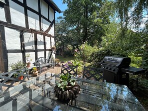 Een terras met een glazen tafel, een barbecue en een vakwerkhuis op de achtergrond.