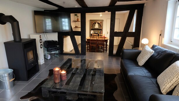 A cozy living room with a fireplace, a TV, a wooden coffee table with candles, a black leather sofa, and a dining area in the background. - Apartment 'Haus Auenland' with Shared Terrace, Garden, and Wi-Fi (Feldatal)