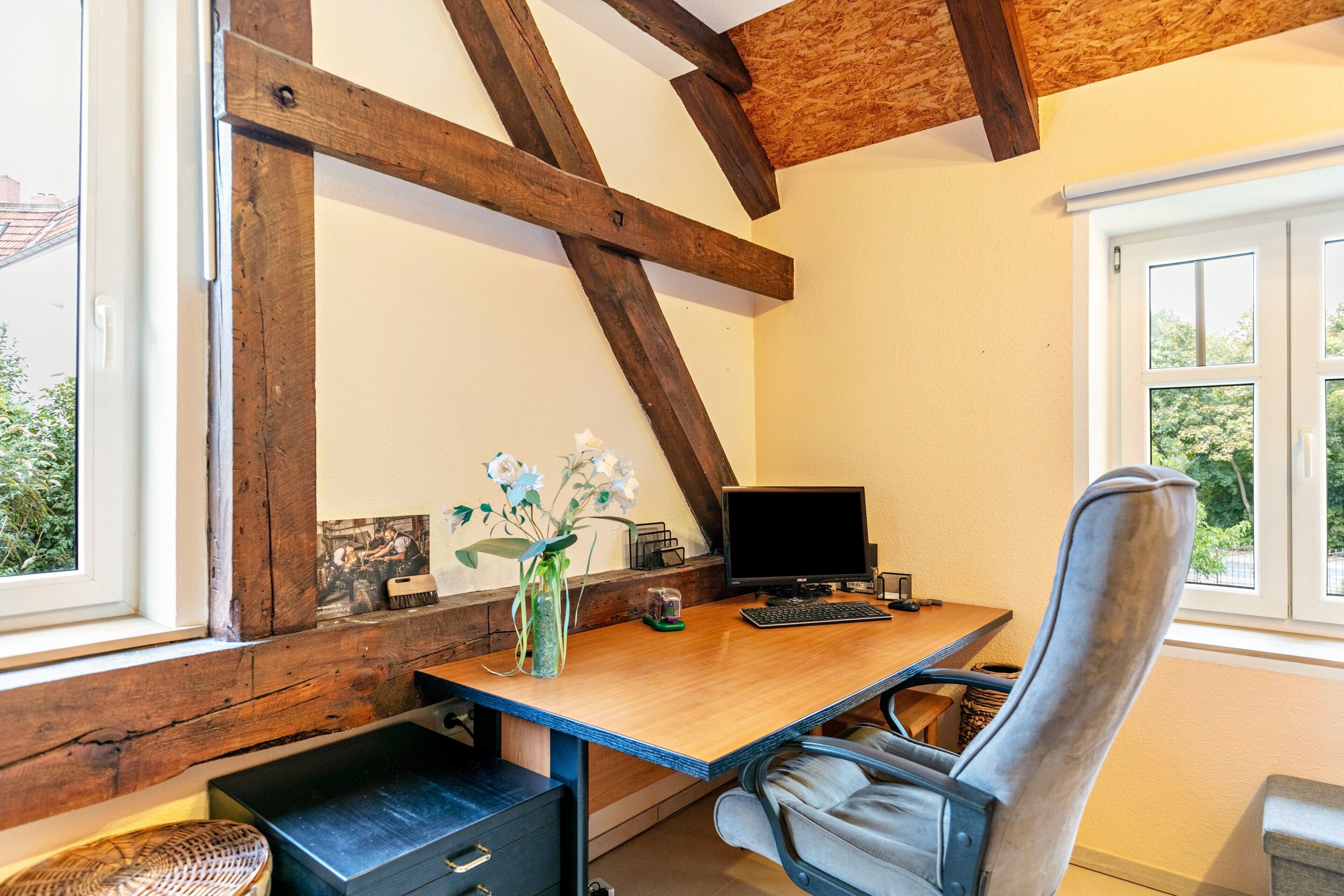 A room with a wooden desk, a computer, a chair, and a window with a view of greenery.