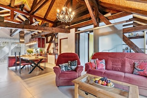 A spacious living area with a high ceiling, exposed wooden beams, a red sofa, a dining table with chairs, and a chandelier.
